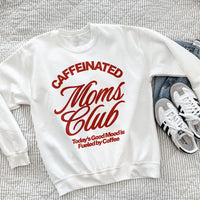 Caffeinated Moms Club white sweatshirt with bold red retro text reading “Today’s Good Mood Is Fueled by Coffee,” styled on a bed with jeans and sneakers.