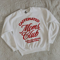 White Caffeinated Moms Club sweatshirt folded neatly on a cozy bed, showing red retro lettering for coffee-loving moms.