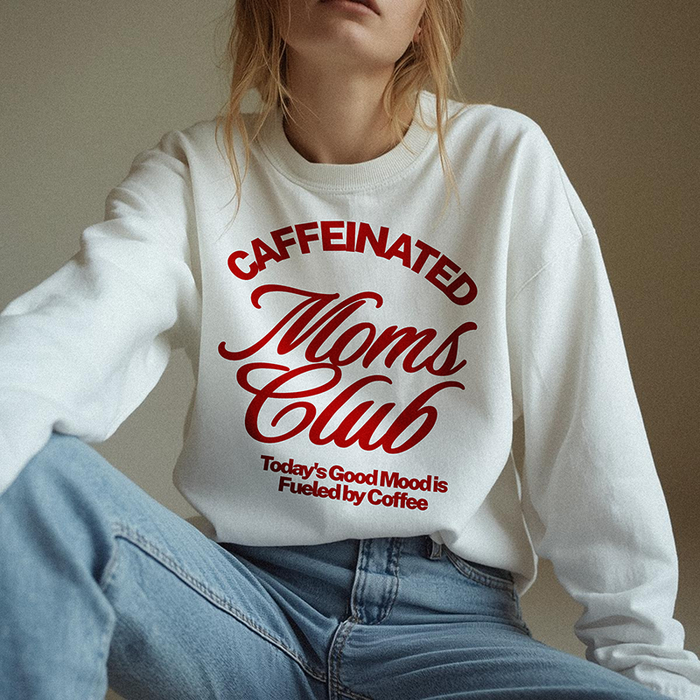Mom wearing white Caffeinated Moms Club crewneck sweatshirt with red print, styled casually with jeans for a cozy, caffeine-fueled morning look.