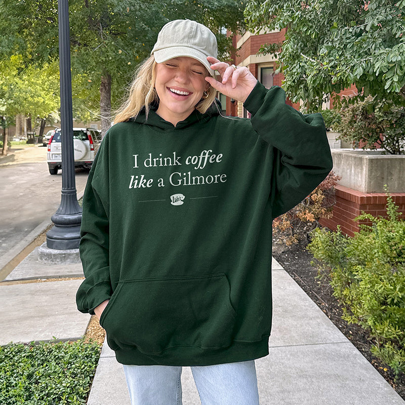Woman walking through neighborhood wearing forest green “I Drink Coffee Like a Gilmore” hoodie with jeans and baseball cap – cozy Gilmore Girls fan look