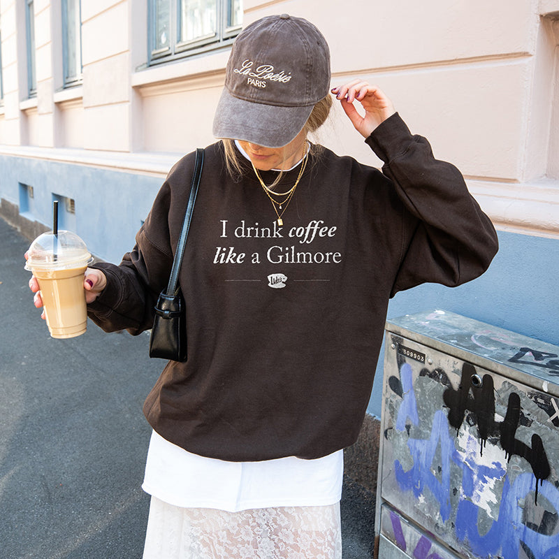 Woman walking with coffee wearing dark chocolate “I Drink Coffee Like a Gilmore” sweatshirt, baseball cap, and skirt – cozy Gilmore Girls fan outfit