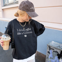 Woman walking with coffee wearing black “I Drink Coffee Like a Gilmore” sweatshirt, baseball cap, and skirt – casual Gilmore Girls inspired outfit