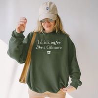 Indoor photo of woman wearing military green “I Drink Coffee Like a Gilmore” sweatshirt styled with skirt – cozy Stars Hollow vibes