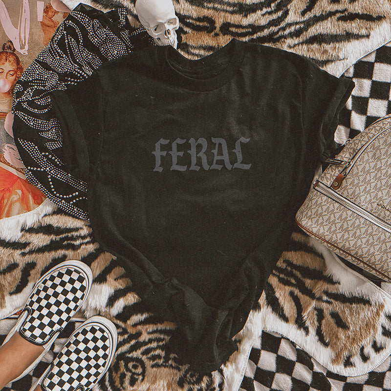 Flatlay of Feral Graphic Tee in black showing old English graphic and garment-dyed texture.