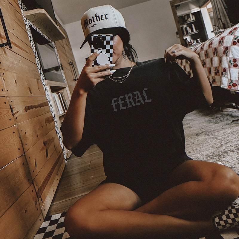 Model wearing Feral Graphic Tee in black with a trucker hat, styled with a casual, edgy look.