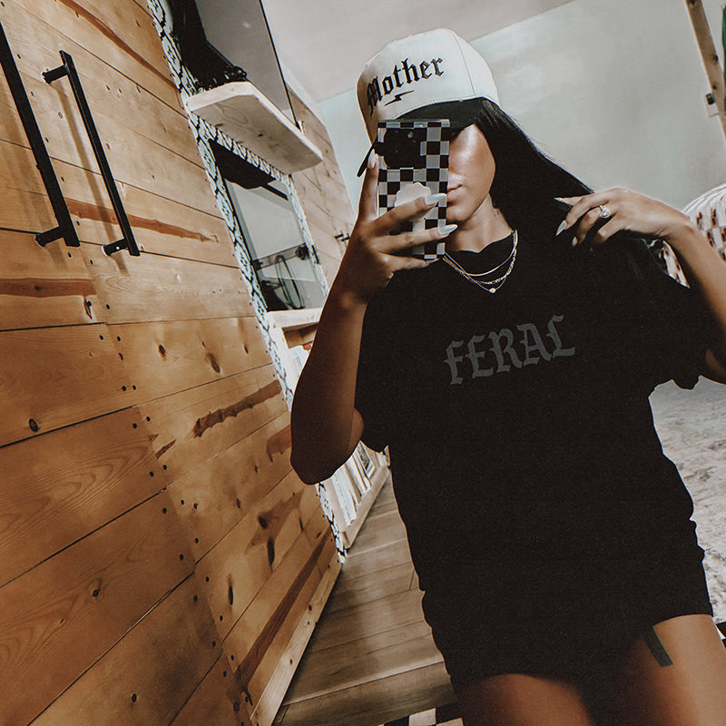 Model lounging in black Feral Graphic Tee with relaxed fit and muted grey old English lettering.