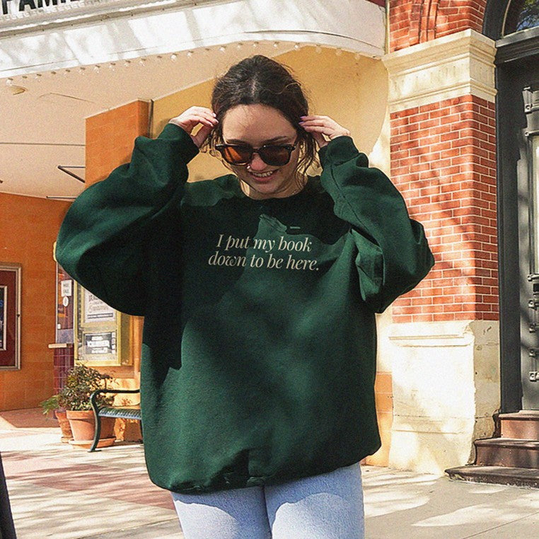 Woman wearing the I Put My Book Down To Be Here Sweatshirt in forest green with jeans and sunglasses – funny book lover crewneck.