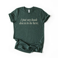 Flatlay of the I Put My Book Down To Be Here Tee in heather forest – witty reading quote shirt for book lovers.