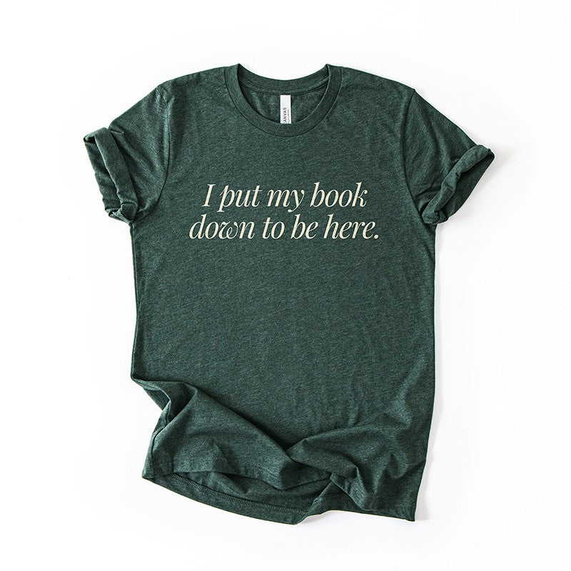Flatlay of the I Put My Book Down To Be Here Tee in heather forest – witty reading quote shirt for book lovers.