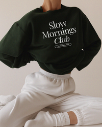 Woman wearing forest green Slow Mornings Club sweatshirt with white sweatpants for a cozy lounge look