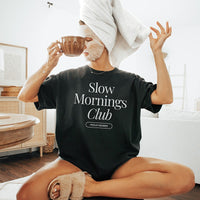 Woman in face mask and hair towel, wearing slippers and holding a cup of coffee while lounging in the black Slow Mornings Club Tee – cozy self-care morning vibes